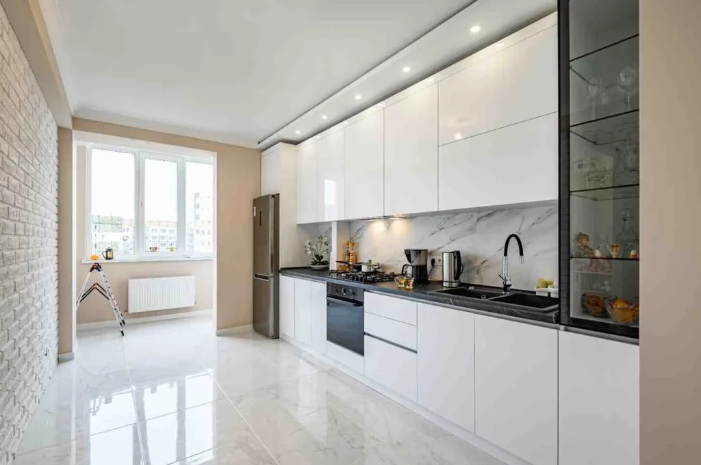 one wall kitchen ideas featuring a modern white gloss kitchen with sleek handleless cabinets, marble-effect backsplash, black countertop, built-in appliances, recessed ceiling lights, and a bright open layout.